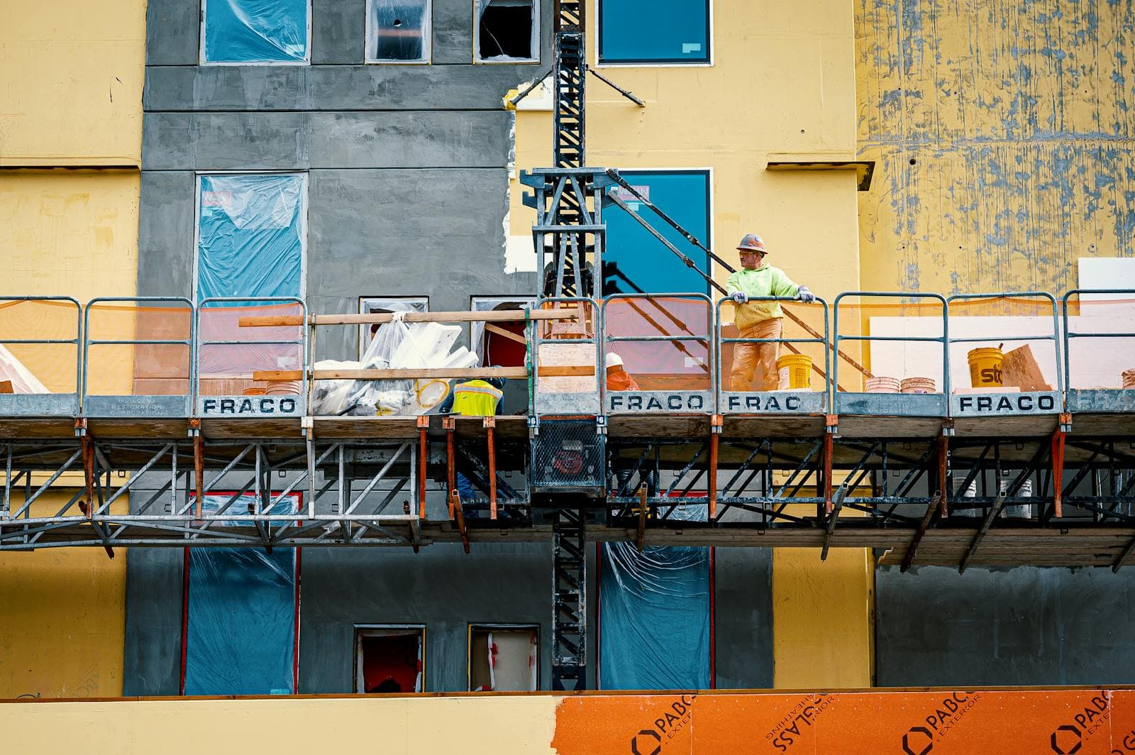 Construction worker in a hard hat and yellow jacket on an elevated work platform against a building facade