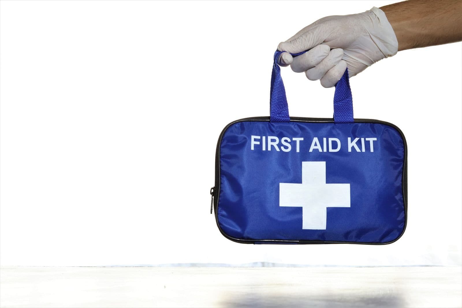 En hånd med hvit hanske holder en blå førstehjelpsveske merket FIRST AID KIT med hvitt kors